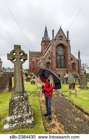 Stock Image - "Exterior view of St. Magnus Cathedral with woman with umbrella in the city of Kirkwall, Mainland Island, Orkney Archipelago, Scotland. ". Fotosearch