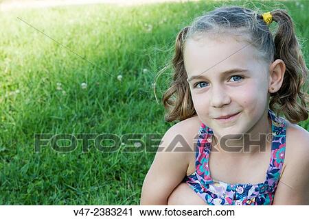 Stock Image - Girl sitting on the grass smiles wing camara. Fotosearch