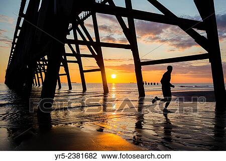 Stock Image - "Jogger at sunrise under Steetley pier on The Headland, Hartlepool, north east England. ". Fotosearch