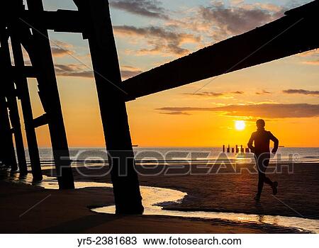 Stock Image - "Jogger at sunrise under Steetley pier on The Headland, Hartlepool, north east England. ". Fotosearch
