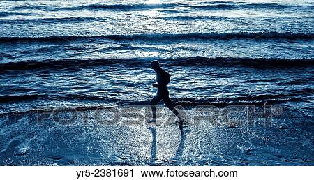 Stock Image - "Man running barefoot along shoreline at sunrise in the North Sea at Seaton Carew, North east England, United Kingdom. ". Fotosearch