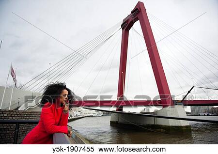 Stock Photography - "Rotterdam, Netherlands. Young, African woman with a red winter coat, pondering and looking out over the river, next to the Willemsbrug in Rotterdam. ". Fotosearch