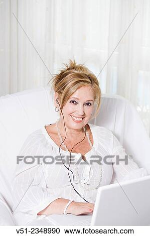 Stock Image - "Senior woman on the couch with laptop, listening to music and smiling at camera". Fotosearch