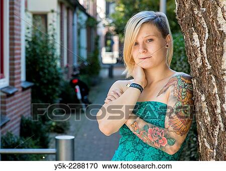 "Tilburg, Netherlands. Young blonde woman with a tattoo on her left arm leaning against a tree in a domestic street. " View Large Photo Image Stock Image - "Tilburg, Netherlands. Young blonde woman with a tattoo on her left arm leaning against a tree in a domestic street. ". Fotosearch