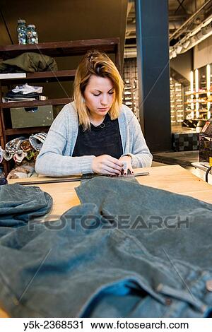 Stock Image - "Tilburg, Netherlands. Young needlewoman buzzy shortening a jeans at her workstation in a fashion store for one of her customers. ". Fotosearch