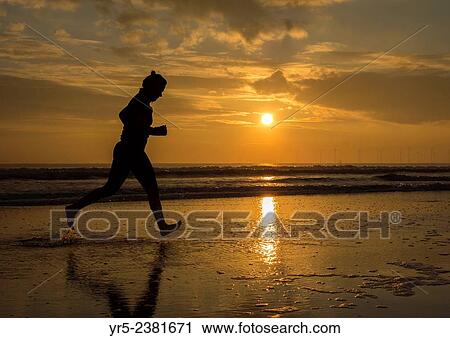 Stock Image - "Woman running barefoot along shoreline at sunrise in the North Sea at Seaton Carew, North east England, United Kingdom. ". Fotosearch