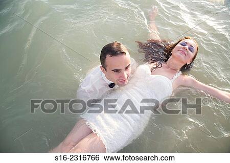 "Young man dressed up in a party suit, holding afloat in shalow sea a young woman in a white party dress. " View Large Photo Image Stock Photograph - "Young man dressed up in a party suit, holding afloat in shalow sea a young woman in a white party dress. ". Fotosearch
