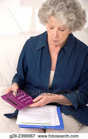 Stock Photo - Beautiful senior woman holding a pocket calculator and reading papers at home. Fotosearch