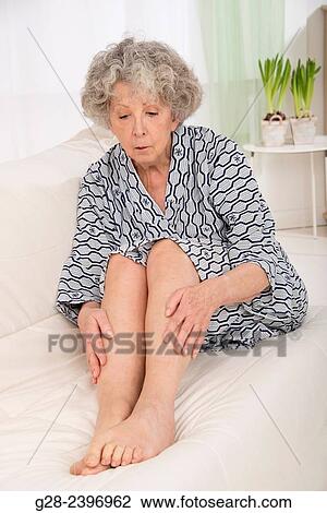 Stock Image - "Beautiful senior woman holding her suffering legs, sitting on a sofa at home with plants in the background". Fotosearch