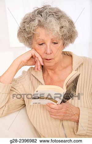 Stock Image - "Beautiful senior woman reading a book, leaning on her elbow and sitting on her sofa at home". Fotosearch