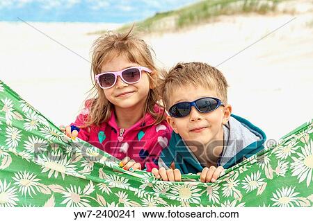 Stock Image - Bother and sister wearing sunglasses behind windbreaker on a beach.. Fotosearch
