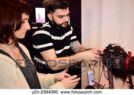 Stock Image - Student hairstylists working on rope braiding hair in a salon while model check her phone.. Fotosearch