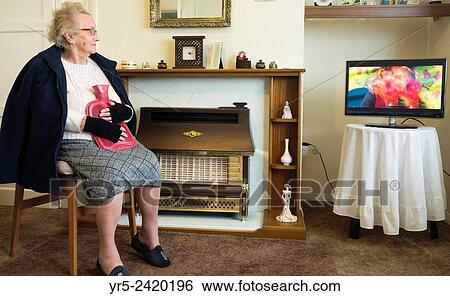 Stock Photograph - 90 year old lady saving fuel by wearing coat and gloves and holding hot water bottle while watching TV in her own home. Billingham, England, UK.. Fotosearch