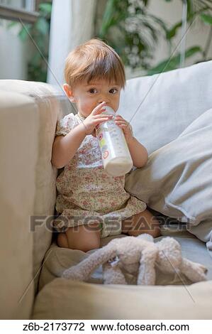 Stock Image - Baby girl, 10 Months Old, Feeding-Bottle, Milk, Indoors, Couch, Window, Plants, Nappy, Stuffed-Toy. Fotosearch