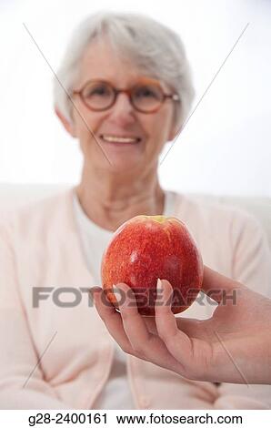 Beautiful senior woman with glasses, blurred in the background, and in the foreground a red apple is offered to her View Large Photo Image Stock Image - Beautiful senior woman with glasses, blurred in the background, and in the foreground a red apple is offered to her. Fotosearch