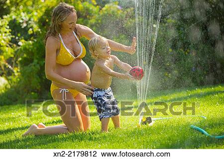 Stock Image - Pregnant woman in yellow bikini plays in the lawn sprinkler with her young son.. Fotosearch