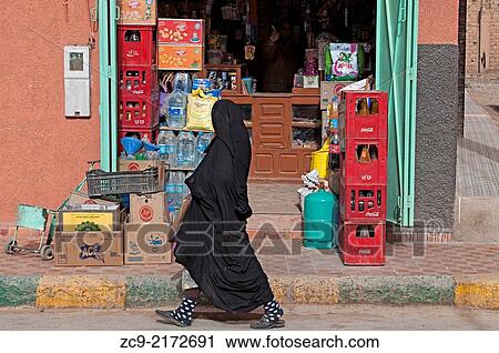 Stock Image - Woman wearing moroccan niqab walking by a shop on the streets of Rissani, Ziz Valley, Morocco.. Fotosearch