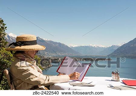 Woman with straw hat sitting in an outdoors restaurant and reading the menu with panoramic view over alpine lake Maggiore with snow-capped mountains in a sunny day with blue sky in Ticino, Switzerland. View Large Photo Image Stock Image - Woman with straw hat sitting in an outdoors restaurant and reading the menu with panoramic view over alpine lake Maggiore with snow-capped mountains in a sunny day with blue sky in Ticino, Switzerland.. Fotosearch