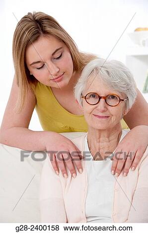 Stock Photo - Young medical help or granddaughter holding beautiful senior woman by her shoulders, and smiling to her, at home. Fotosearch