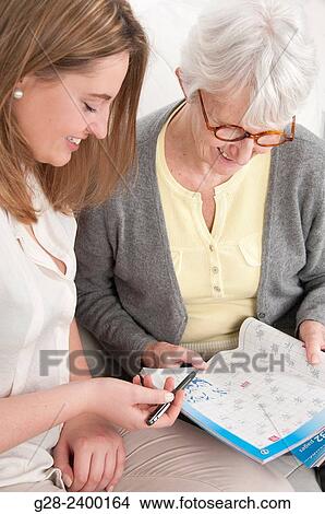 Picture - Young nurse or help or grand daughter holding a pen for patient or grand mother busy making crosswords. Fotosearch