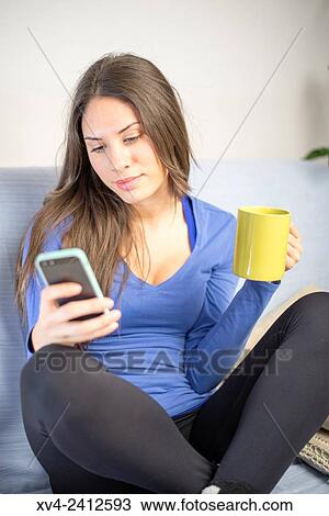 Stock Image - Young Woman chating on her sofa and drinking hot beverage.. Fotosearch