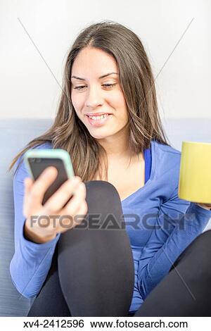Stock Photograph - Young Woman chating on her sofa and drinking hot beverage.. Fotosearch