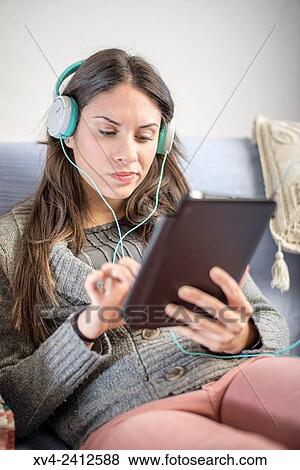 Young Woman relaxing on her sofa and listening to music on her tablet with Headphones. View Large Photo Image Stock Photo - Young Woman relaxing on her sofa and listening to music on her tablet with Headphones.. Fotosearch