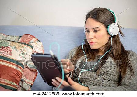 Stock Photo - Young Woman relaxing on her sofa and listening to music on her tablet with Headphones.. Fotosearch