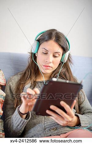 Young Woman relaxing on her sofa and listening to music on her tablet with Headphones. View Large Photo Image Stock Photograph - Young Woman relaxing on her sofa and listening to music on her tablet with Headphones.. Fotosearch