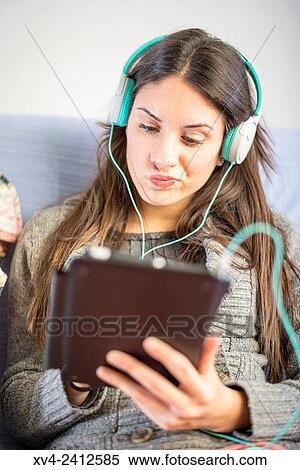 Stock Photography - Young Woman relaxing on her sofa and listening to music on her tablet with Headphones.. Fotosearch