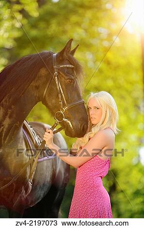 Stock Image - Young woman standing beside a Friesian horse in late sunlight.. Fotosearch