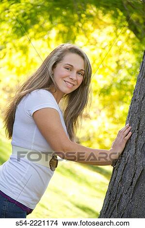 Picture - A happy teen girl leans against a tree at a park in the fall.. Fotosearch