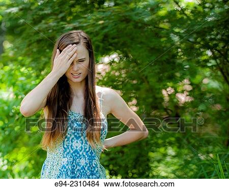 An 18 year old woman holding her hand to her forehead with a grimace on her face, standing in a garden. View Large Photo Image Picture - An 18 year old woman holding her hand to her forehead with a grimace on her face, standing in a garden.. Fotosearch