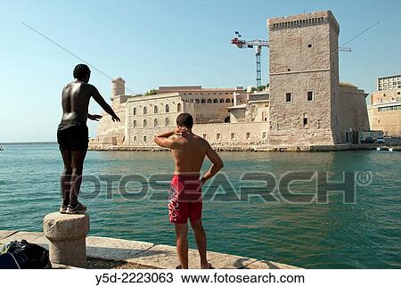 Stock Image - boys jumping to sea entrance to old harborin Marseille, Fort St-Jean in the background, Marseille, France.. Fotosearch