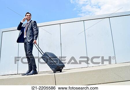 Businessman, standing, talking with a mobile while holding a suitcase on wheels outdoors. View Large Photo Image Stock Photograph - Businessman, standing, talking with a mobile while holding a suitcase on wheels outdoors.. Fotosearch