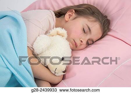 Stock Image - Little girl asleep in bed cuddling her teddy bear. Fotosearch