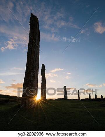 Stock Photograph - Setting sun shines behind standing stone at Ring of Brodgar, Orkney, Scotland.. Fotosearch