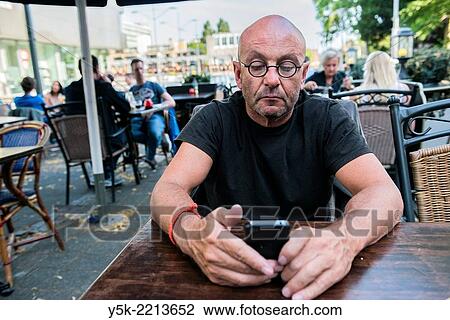 Tilburg, Netherlands. Bald, be glassed and middle-aged man, reading his iPhone messages, while sitting down at an outdoor restaurant table, waiting for his diner. View Large Photo Image Stock Image - Tilburg, Netherlands. Bald, be glassed and middle-aged man, reading his iPhone messages, while sitting down at an outdoor restaurant table, waiting for his diner.. Fotosearch