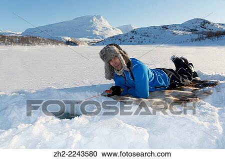 Stock Image - White man, 40-45 years old, icefishing, lying on a reindeer fur in Kiruna, Riksgränsen, Swedish lapland.. Fotosearch