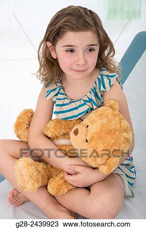 Stock Image - Worried and crying little girl sitting on a medical couch holding her teddy bear. Fotosearch