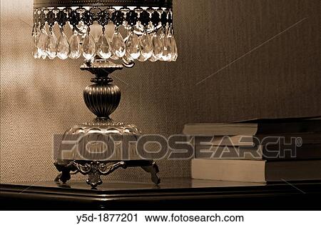 Stock Image - Crystal antique lamp on the shelf, light on, next to laying books, symbol of home seclusion and privacy. Fotosearch