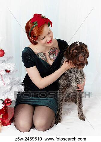 Stock Image - Happy pregnant woman and dog sit together alongside the christmas tree.. Fotosearch