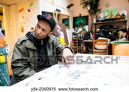 Stock Photography - Rotterdam, Netherlands. North African artist, wearing a winter coat and cap, working on a sketch on the main production table of Via Kunst StudioÂ´s.. Fotosearch