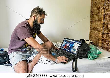 Tilburg, Netherlands. Brazilian male traveller sitting on the bed of his Airbnb guest room with his laptop computer at arms length. View Large Photo Image Stock Photo - Tilburg, Netherlands. Brazilian male traveller sitting on the bed of his Airbnb guest room with his laptop computer at arms length.. Fotosearch
