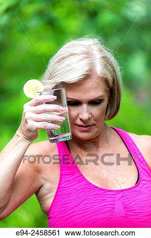 Stock Image - 56 year old blond woman holding a glass of water to her head to cool off.. Fotosearch