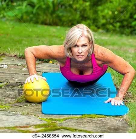A 56 year old blond woman doing a push up on a yoga mat and balancing on a ball on one hand, outdoors. View Large Photo Image Picture - A 56 year old blond woman doing a push up on a yoga mat and balancing on a ball on one hand, outdoors.. Fotosearch