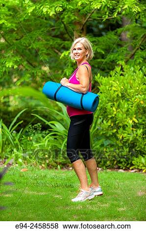 A 56 year old blond woman dressed in fitness clothing and holding a yoga mat, smiling at the camera, outdoors. View Large Photo Image Stock Photo - A 56 year old blond woman dressed in fitness clothing and holding a yoga mat, smiling at the camera, outdoors.. Fotosearch