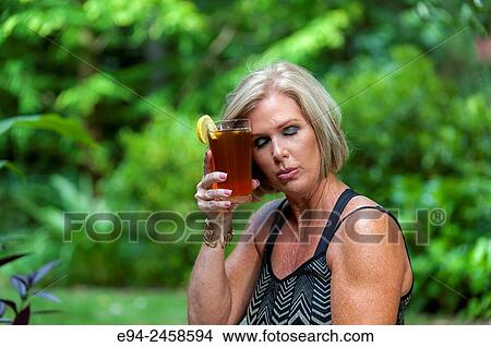 A 56 year old blond woman holding a glass of iced tea holding it to her forehead with her eyes closed. View Large Photo Image Picture - A 56 year old blond woman holding a glass of iced tea holding it to her forehead with her eyes closed.. Fotosearch
