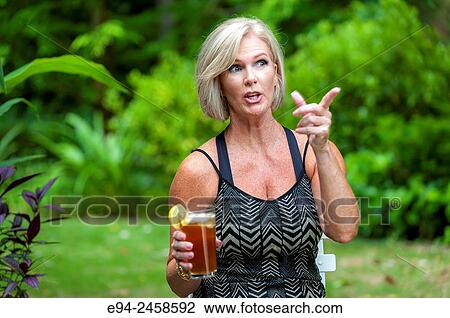 A 56 year old blond woman holding a glass of iced tea pointing and looking away from the camera, outdoors. View Large Photo Image Stock Image - A 56 year old blond woman holding a glass of iced tea pointing and looking away from the camera, outdoors.. Fotosearch