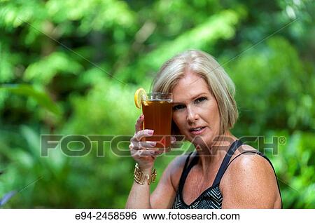 A 56 year old blond woman holding a glass of iced tea holding it to her forehead looking at the camera. View Large Photo Image Stock Photograph - A 56 year old blond woman holding a glass of iced tea holding it to her forehead looking at the camera.. Fotosearch